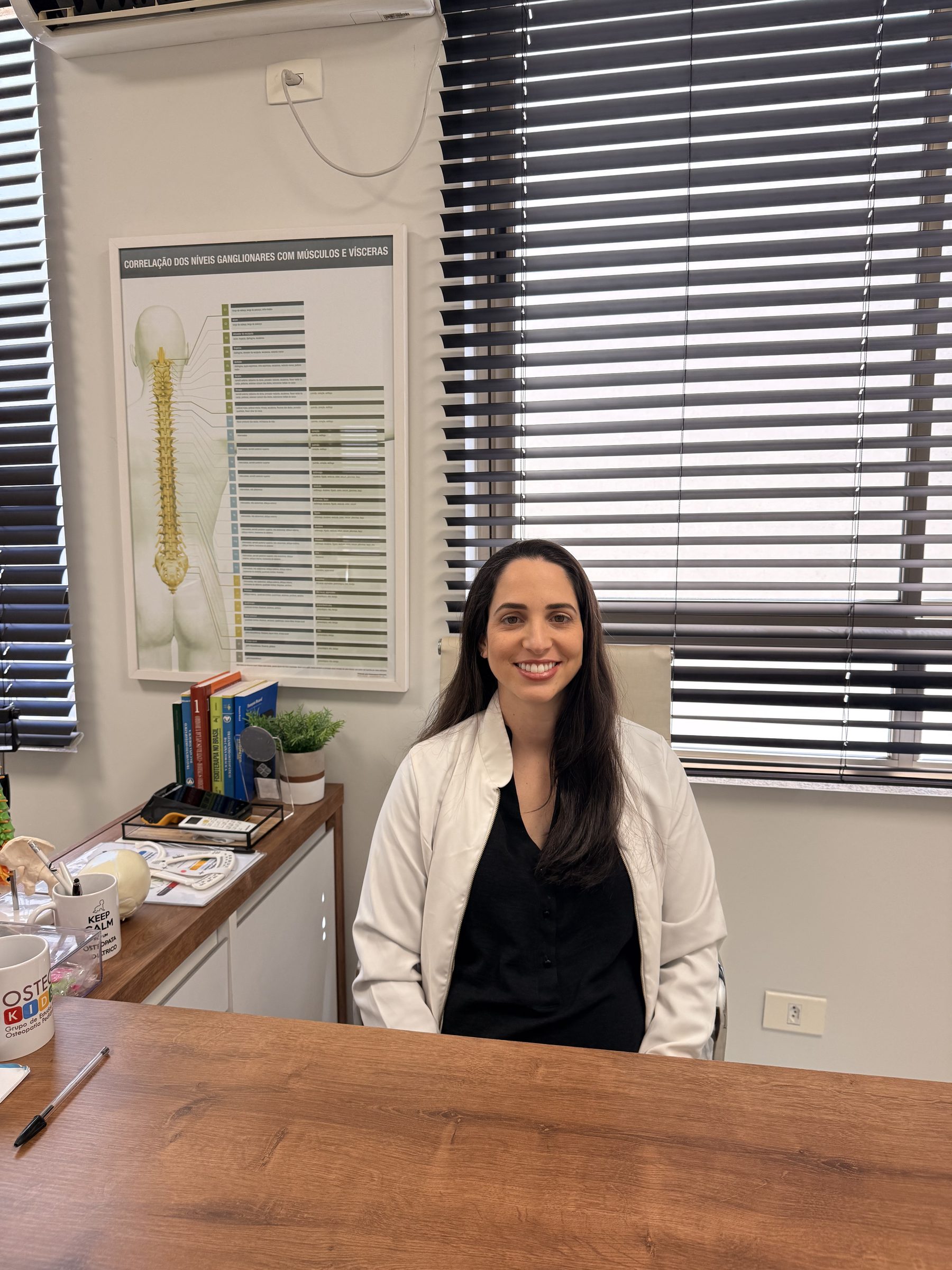 Paula à mesa do consultório, sorrindo para quem chega